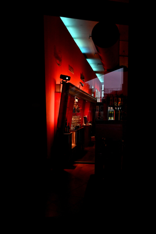A dimly lit interior space, possibly a bar or lounge, with a strong red light illuminating a wall on the left. A large framed mirror or artwork leans against the red wall, reflecting glassware. A strip of blue light is visible on the ceiling, contrasting with the red. The overall atmosphere is dark and moody.