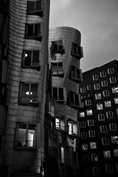Black and white image of the distinctive, curved Gehry Buildings in Düsseldorf's MedienHafen, featuring their metallic facades and illuminated windows under an overcast sky."