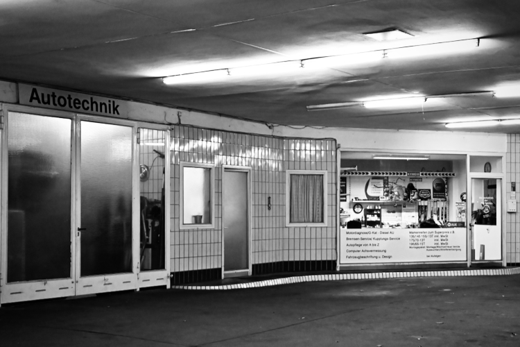 A black and white photo of an 'Autotechnik' garage or workshop entrance with large glass doors and tiled walls. Various signs and posters are visible inside the right-hand section, under fluorescent lighting.