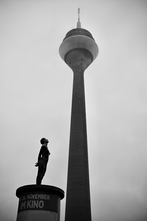 Black and white photo of a statue of a person looking upwards at a very tall, slender telecommunications tower. The statue stands on a cylindrical pedestal that has text partially visible, including '26. NOVEMBER IM KINO'.