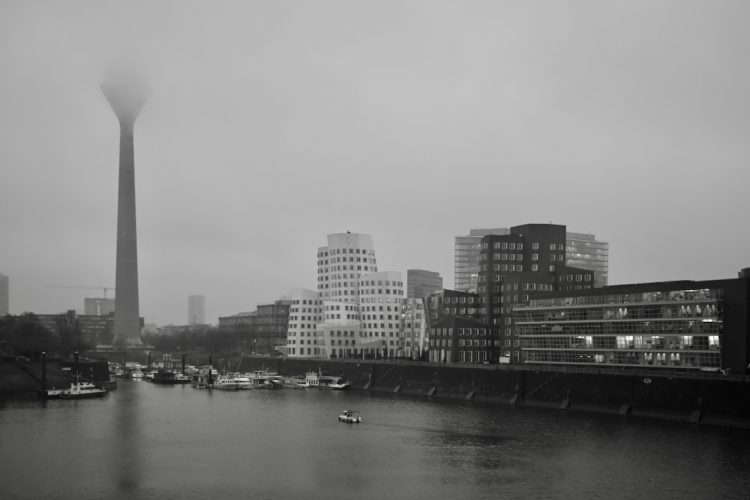 A foggy black-and-white cityscape featuring a tall tower partially shrouded in mist. Boats are docked along the riverbank, with modern buildings lining the waterfront under an overcast sky.
