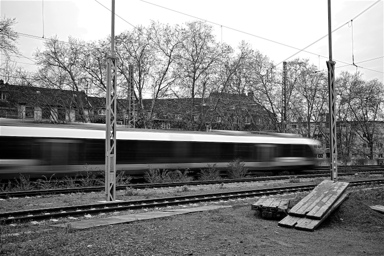 A black and white image capturing a blurred train speeding past on tracks, with bare trees and buildings in the background. Utility poles stand beside the tracks, and some wooden planks are on the ground in the foreground.