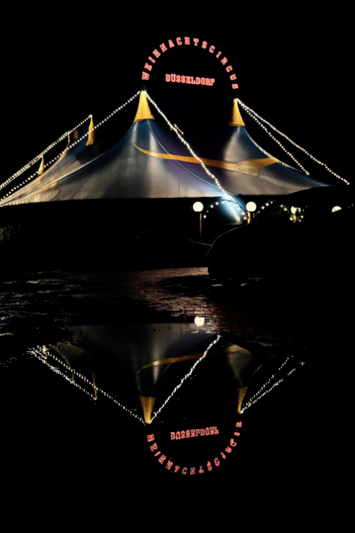 A brightly lit circus tent at night, with strings of lights outlining its peaks and sides. The words 'WEIHNACHTSCIRKUS' and 'DÜSSELDORF' are spelled out in lights on the tent. The tent and its lights are perfectly reflected in a dark, still body of water below, creating a striking symmetrical image.