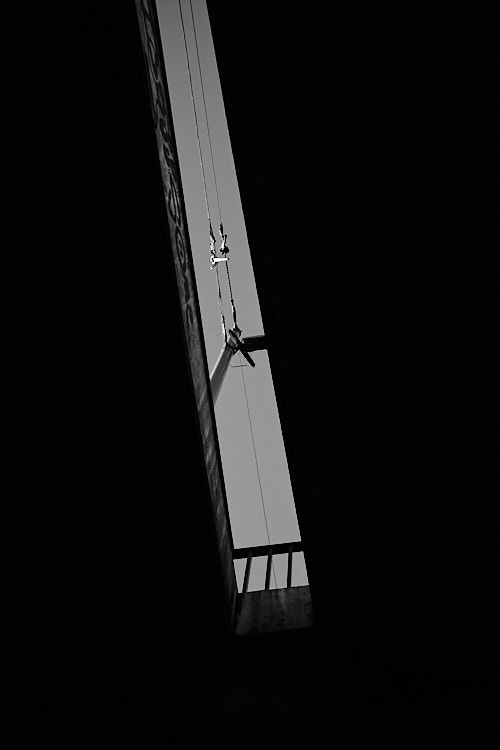 A black and white photo showing a narrow vertical gap between two dark urban structures, revealing a bright sky with a utility pole and intersecting power lines. The composition emphasizes strong contrasts, geometric lines, and the interplay of light and shadow in an urban environment.