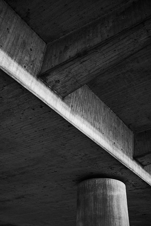 A black and white photo showing the underside of a concrete structure with beams and a cylindrical pillar. The composition emphasizes geometric shapes, textures, and the interplay of light and shadow in urban architecture.