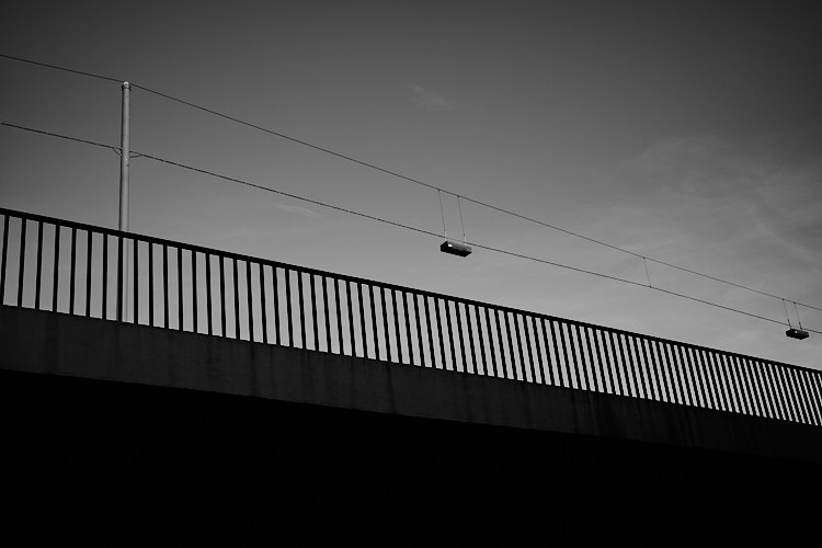 A black and white photo showing a horizontal urban railing silhouetted against a clear sky, with overhead power lines stretching across the frame. The composition emphasizes geometric lines, contrast, and minimalist urban structure.