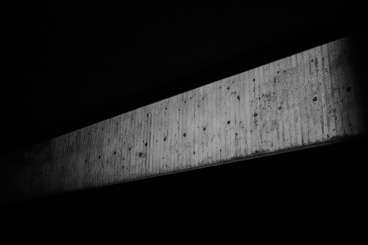 A black and white photo showing a narrow horizontal beam of light illuminating a textured concrete surface, surrounded by deep shadows. The composition highlights contrast, texture, and minimalist urban geometry.