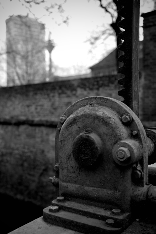 Black-and-white photograph showing rusty mechanical parts of the Düsseldorf Spee'scher Graben water control structure in the foreground with the Mannesmann Tower and Düsseldorf TV Tower blurred in the background.