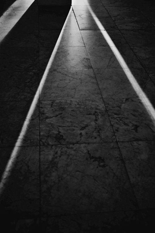 Black and white image of two sharp, diagonal beams of light cutting across a textured tiled floor, featuring a distinct footprint in the illuminated area.