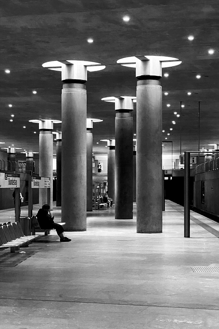 Black and white photo of a modern, minimalist underground station platform, characterized by several large, cylindrical concrete pillars with distinctive mushroom-shaped light fixtures on top. A person is sitting on a bench on the left side of the platform, and a sign partially reading 'Bundestag' is visible nearby. The ceiling is dotted with numerous small lights, and the platform appears clean and spacious.