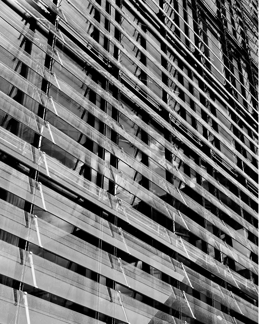 Black and white close-up photo of a modern building facade, featuring numerous horizontal slats or louvers that create a strong linear pattern. Reflections of the sky and possibly other structures are visible in the glass behind the slats, adding depth to the geometric design.