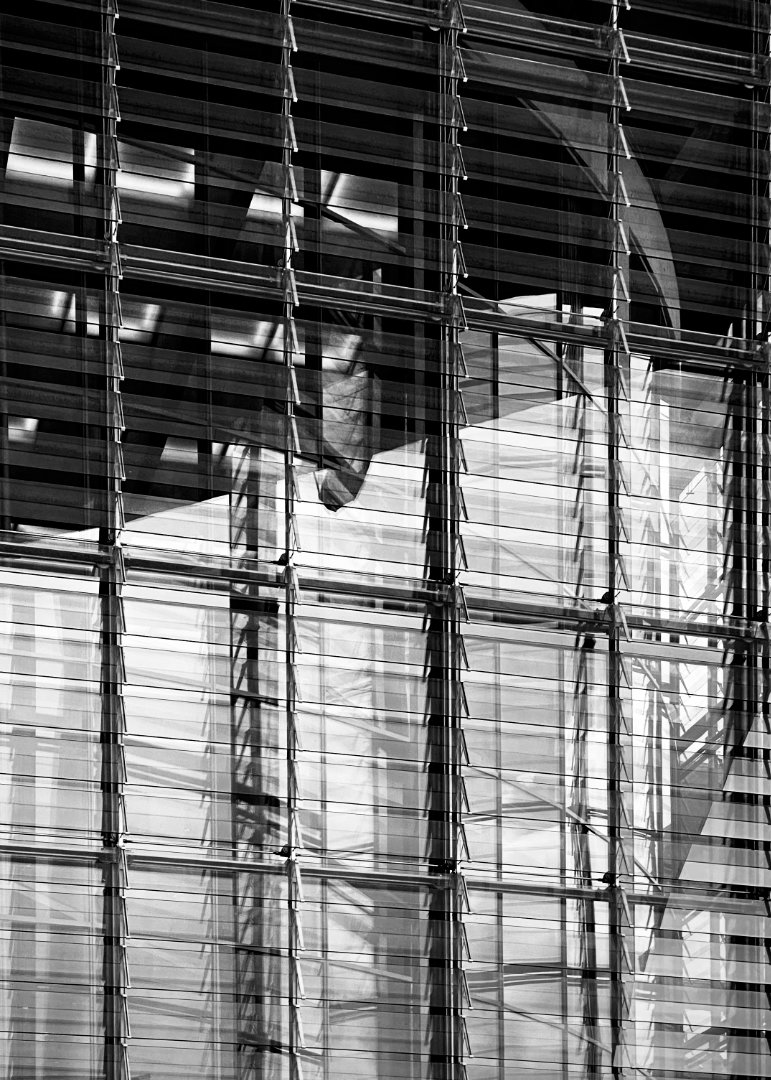 Black and white close-up of a modern building facade, characterized by a grid of horizontal slats or louvers over a glass surface. Complex reflections and shadows of internal structures or surrounding elements are visible through the slats, creating a dynamic and abstract geometric pattern.
