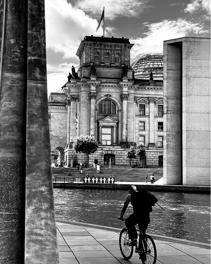 Black and white photo of a person on a bicycle riding along a waterfront promenade, with their back to the viewer. In the background, the grand, classical Reichstag building with a flag flying and a dome is prominently featured, with people visible on its steps. To the right of the Reichstag, a modern, stark concrete building stands. Two large, textured vertical columns partially frame the scene on the left.