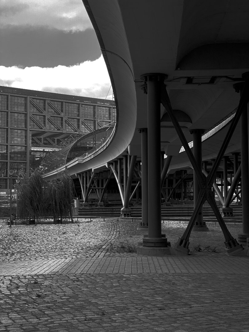 Black and white photo looking up at the underside of a modern, curved elevated walkway or bridge, supported by numerous pillars and diagonal beams. A large, contemporary building with a grid-like facade is visible in the background to the left, under a sky with a dramatic cloud. The ground below is paved with cobblestones, and sunlight casts shadows.