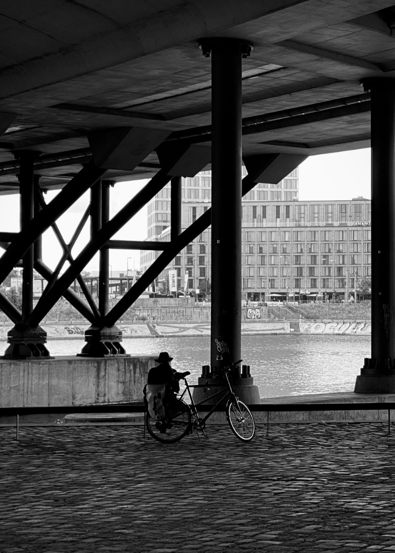 Black and white photo taken from under a large bridge, showing a person sitting on a bench with a bicycle next to them, looking out at a river. The bridge's concrete and metal support structures are prominent in the foreground, and modern buildings are visible on the far bank of the river.