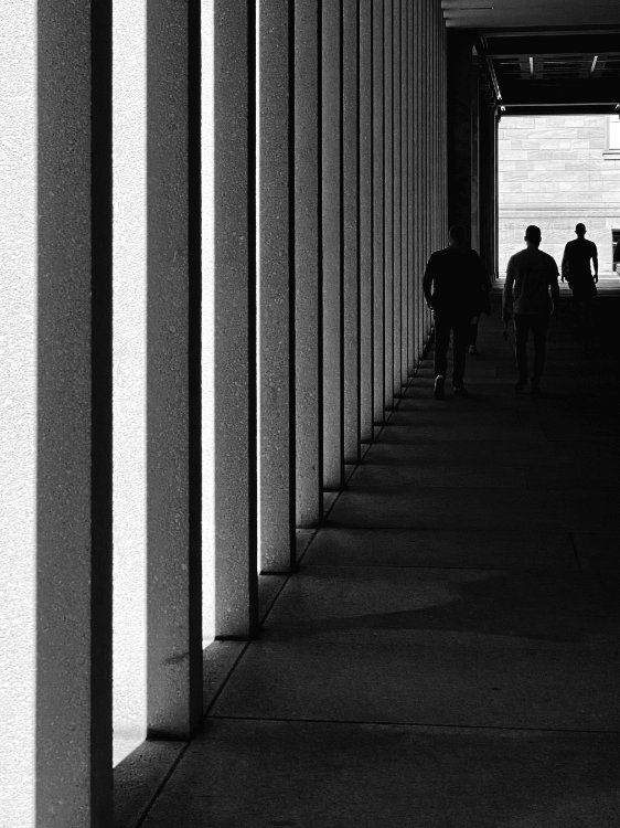 Black-and-white photo of a dark corridor lined with vertical columns creating strong light and shadow patterns. Three shadowy, silhouetted figures walk toward a brightly lit exit at the end of the passage, creating a mysterious atmosphere.