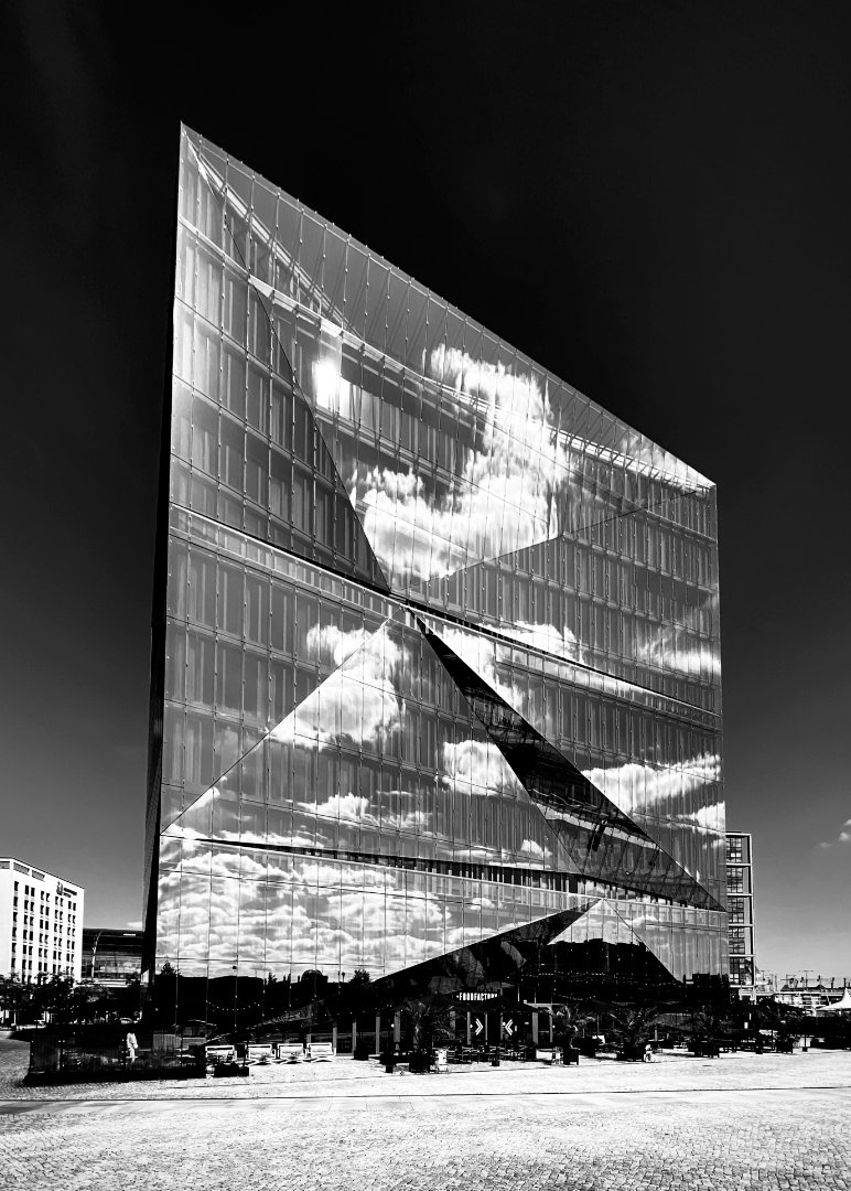 Black and white photo of a striking modern glass building with an angular, faceted design. The building's reflective surface perfectly mirrors dramatic clouds in the sky, creating a dynamic interplay of light and shadow.