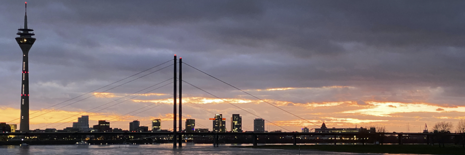 Panoramic view of a city skyline at sunset or sunrise, featuring a tall telecommunications tower, a cable-stayed bridge over the river Rhine, and illuminated buildings. The sky is filled with dramatic clouds displaying vibrant orange and yellow hues.