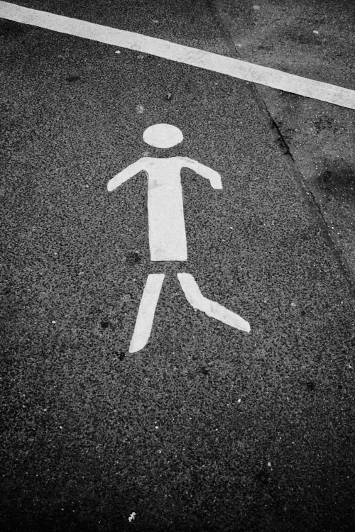 White pedestrian symbol painted on textured asphalt with a diagonal white line crossing the top part of the image, captured in black and white.