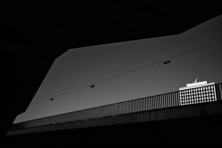Black and white photo of an urban scene featuring a silhouetted bridge railing and overhead cables against a clear sky, with a brightly lit modern high-rise building in the background.