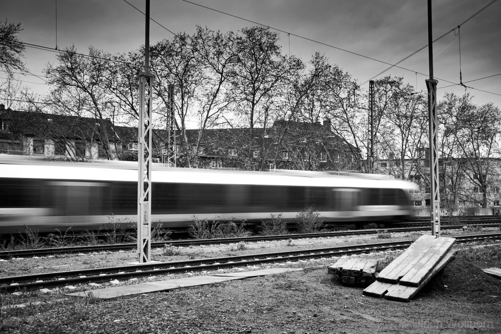 Black and white image of a blurred train moving along railway tracks in an urban area with leafless trees and residential buildings in the background.