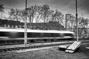 Black and white image of a blurred train moving along railway tracks in an urban area with leafless trees and residential buildings in the background.