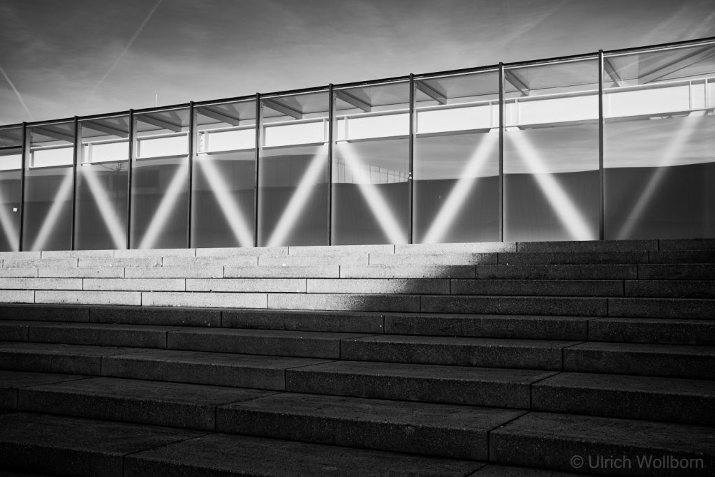 Black and white photo showing a series of glass panels with diagonal light reflections creating a repeating V-shaped pattern above wide concrete steps under a clear sky.