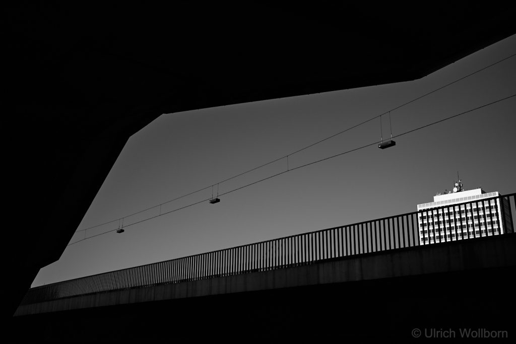Black and white photo of an urban scene featuring a silhouetted bridge railing and overhead cables against a clear sky, with a brightly lit modern high-rise building in the background.