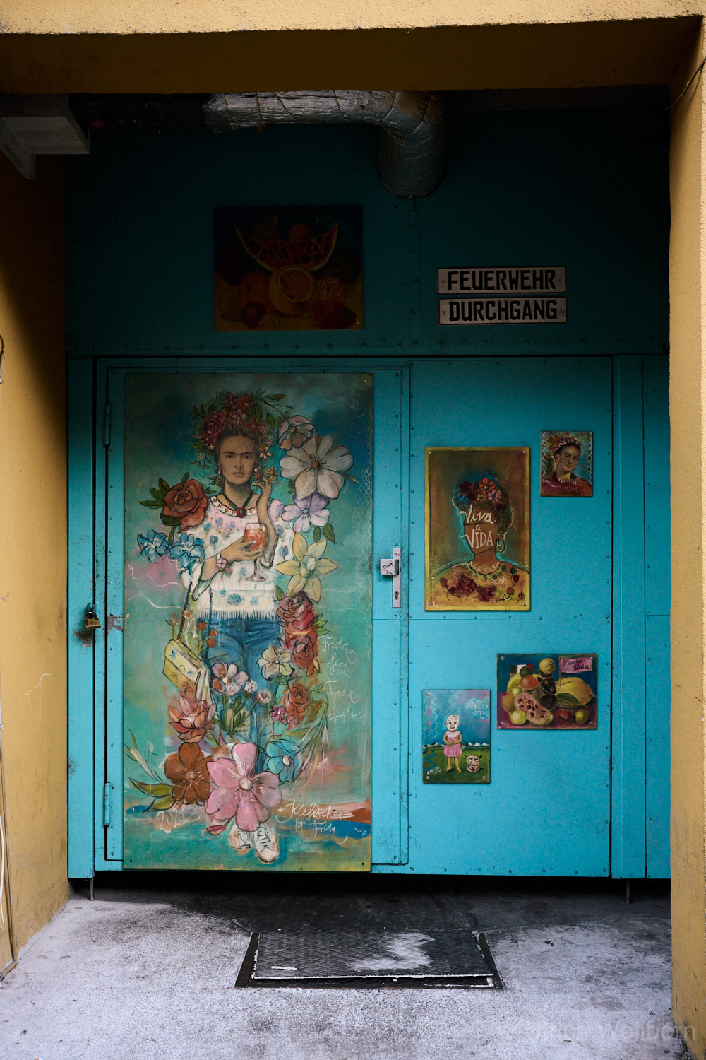 Blue metal door decorated with vibrant paintings of Frida Kahlo and floral motifs, partially open to reveal a dimly lit industrial passageway with exposed pipes and stacked crates. The sign above the door reads "Feuerwehr Durchgang" (Fire Department Passage).