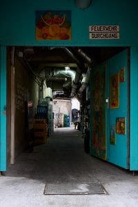 Blue metal door decorated with vibrant paintings of Frida Kahlo and floral motifs, partially open to reveal a dimly lit industrial passageway with exposed pipes and stacked crates. The sign above the door reads "Feuerwehr Durchgang" (Fire Department Passage).
