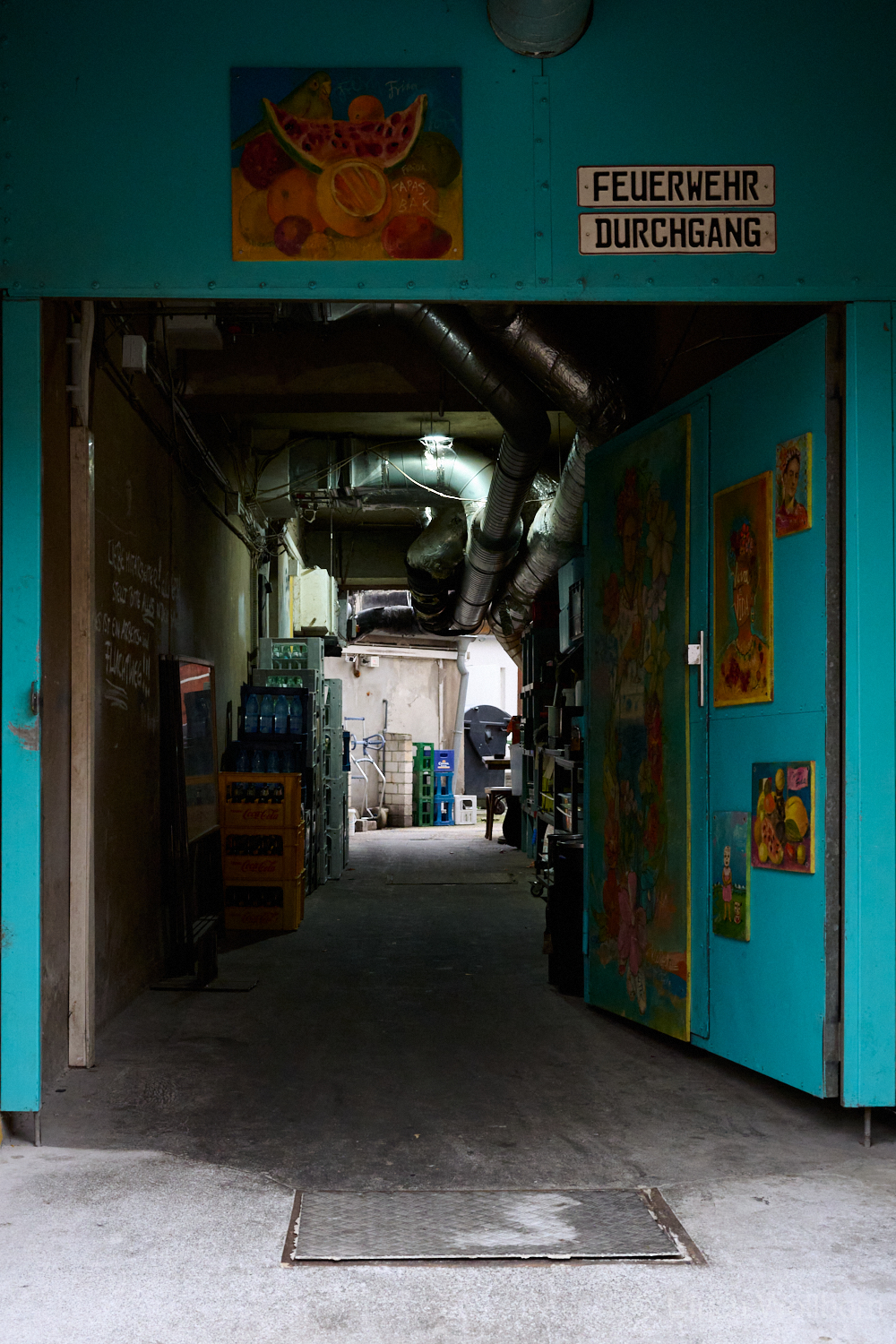 Blue metal door decorated with vibrant paintings of Frida Kahlo and floral motifs, partially open to reveal a dimly lit industrial passageway with exposed pipes and stacked crates. The sign above the door reads "Feuerwehr Durchgang" (Fire Department Passage).