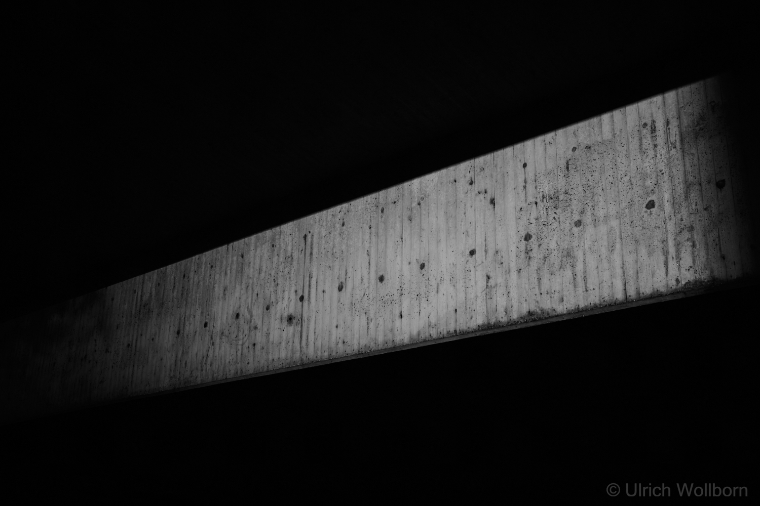 A black and white photo showing a narrow horizontal beam of light illuminating a textured concrete surface, surrounded by deep shadows. The composition highlights contrast, texture, and minimalist urban geometry.