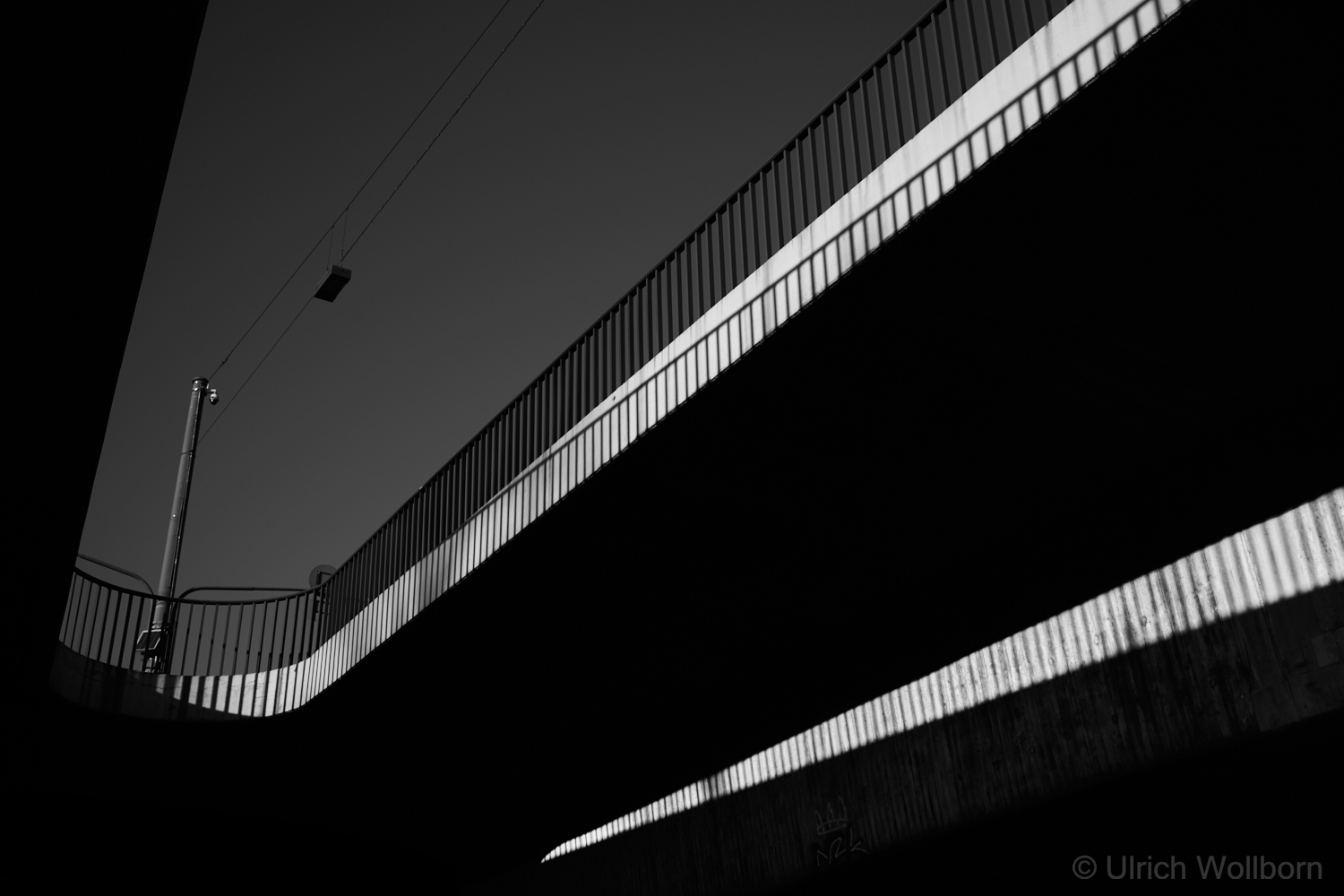 A black and white photo of a bridge taken from below, showing strong diagonal lines created by the bridge's railings and shadows, with a clear sky in the background and a streetlight pole visible on the left side.