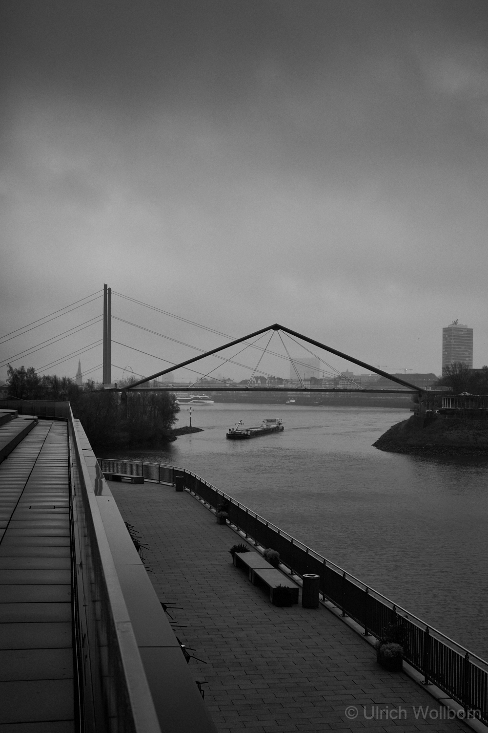 Black and white photo of a foggy river landscape featuring a modern suspension bridge spanning the water, a cargo boat moving along the river, and a paved riverside walkway with benches and planters on a cloudy day.