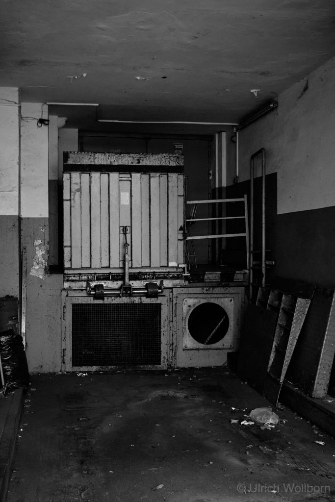 Black and white photo of an industrial loading dock area featuring large metal containers, a grated vent, and scattered debris on the floor in a worn, dimly lit environment.