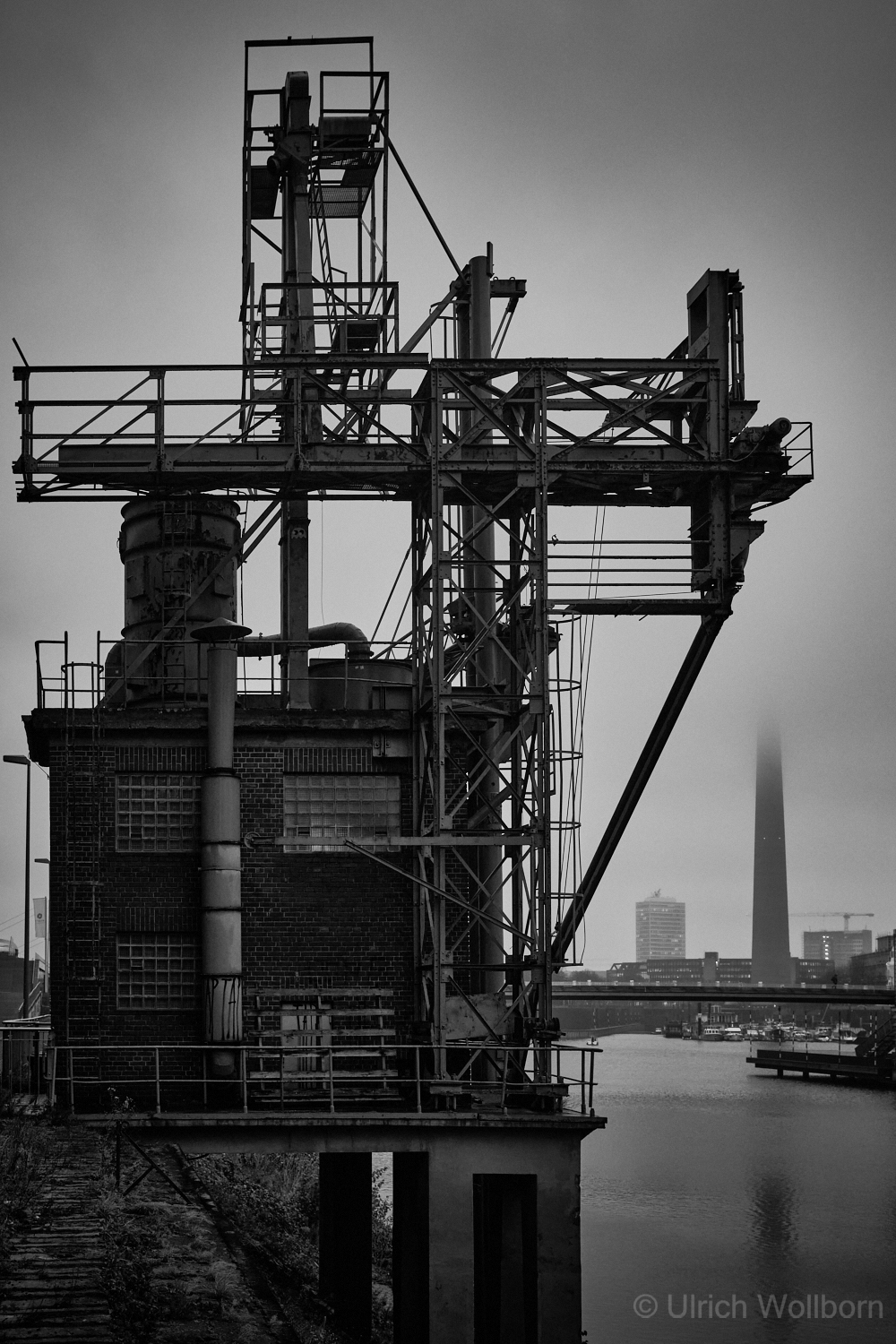 Black and white photo of an old industrial metal structure with pipes and scaffolding beside a river, with a fog-covered city skyline and the silhouette of the Rheinturm tower visible in the background.