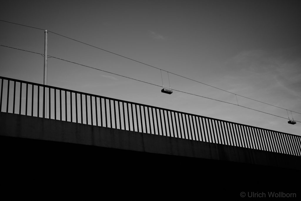 A black and white photo showing a horizontal urban railing silhouetted against a clear sky, with overhead power lines stretching across the frame. The composition emphasizes geometric lines, contrast, and minimalist urban structure.