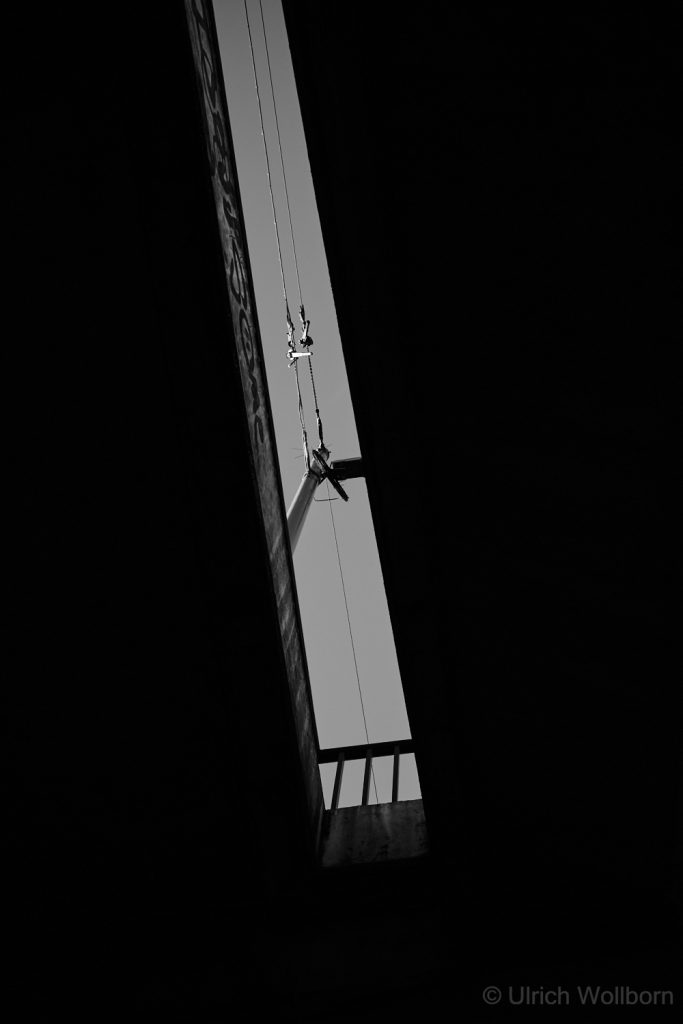 A black and white photo showing a narrow vertical gap between two dark urban structures, revealing a bright sky with a utility pole and intersecting power lines. The composition emphasizes strong contrasts, geometric lines, and the interplay of light and shadow in an urban environment.