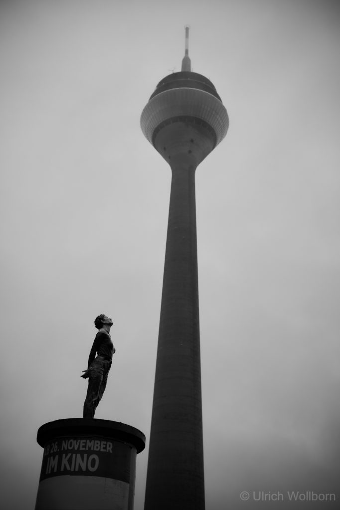 Black and white photo of the statue "Marlies" from the 10 Pillar Saints by artist Christoph Pöggeler, standing on a Litfaß column, looking up towards the Rheinturm tower in Düsseldorf under an overcast sky.