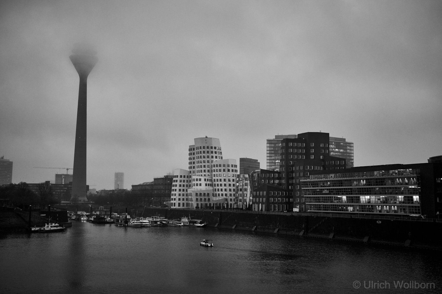 Black and white photo of Düsseldorf’s skyline featuring the Rheinturm tower partially obscured by fog, modern buildings including the iconic Gehry-designed structures, and boats docked along the river on a cloudy day.