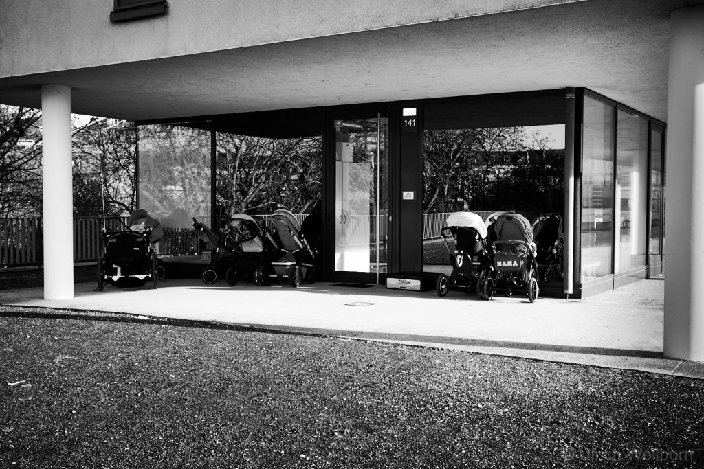 Black and white photo of a modern building entrance with glass walls and white pillars, showing several baby strollers lined up outside on the concrete porch, with reflections of trees visible in the glass.