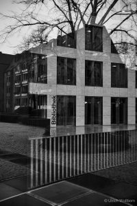 Black and white photo of a modern library building with large windows, reflected in glass showing bare tree branches and surrounding architecture, creating a layered and artistic effect. The word "Bibliothek" is visible on the building facade.