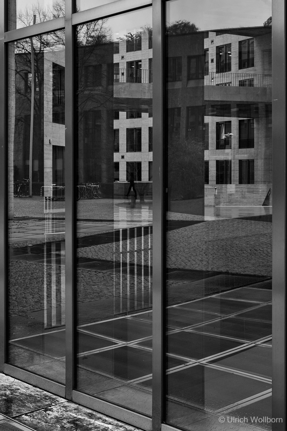 Black and white image showing the reflection of a modern building with large windows and a cobblestone courtyard in tall glass panels. A person is walking in the background, and the scene captures geometric patterns and architectural details through the reflections.