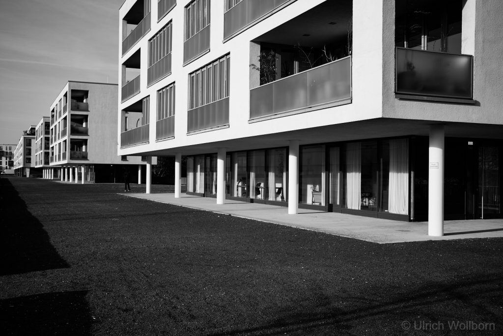 Black and white photo of a row of modern apartment buildings featuring large glass windows and balconies, with white pillars supporting the upper floors and a wide open space in front.