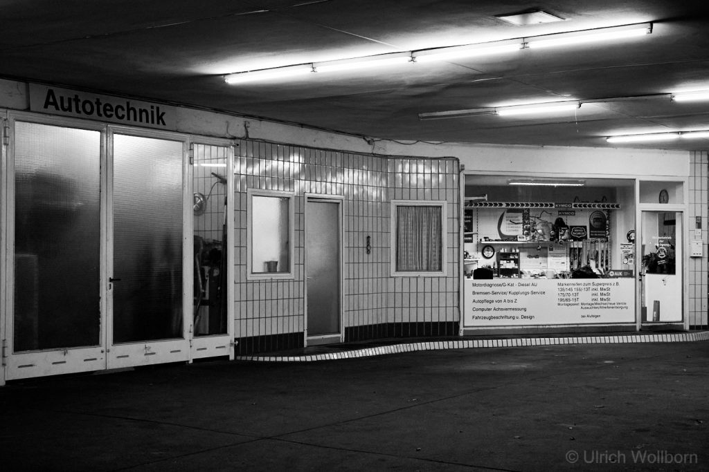 A black and white photo of an 'Autotechnik' garage or workshop entrance with large glass doors and tiled walls. Various signs and posters are visible inside the right-hand section, under fluorescent lighting.