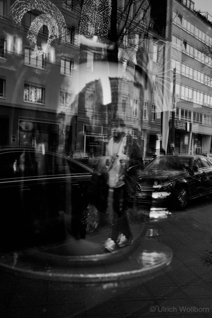 Black and white photo showing the reflection of a person walking on a city street, visible through a shop window glass that also reflects surrounding buildings, cars, and festive lights, creating a layered and ghostly urban scene.
