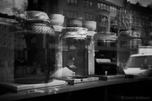 Black and white photo showing large glass jars with cloth covers on a wooden shelf, with the reflection of a person and surrounding buildings visible in the glass window.