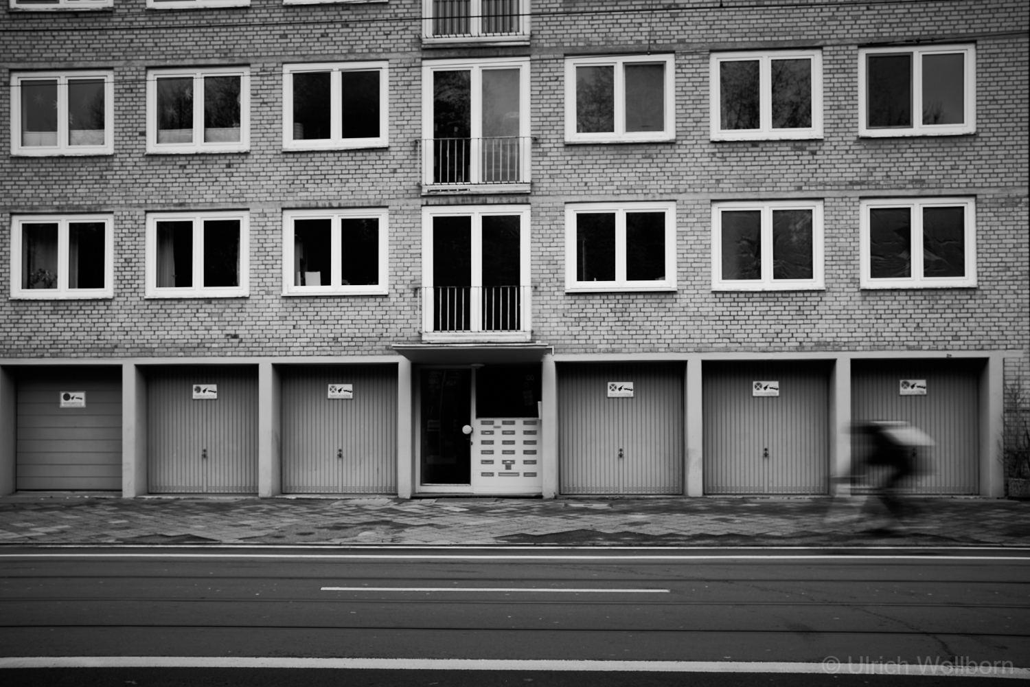 Black-and-white photo of a multi-story brick apartment building with a row of garage doors and a central entrance on the ground floor. Above are multiple windows and small balconies. In the foreground, a street with tram tracks and road markings is visible, with a blurred figure, a cyclist, moving quickly on the right, conveying a sense of urban motion.