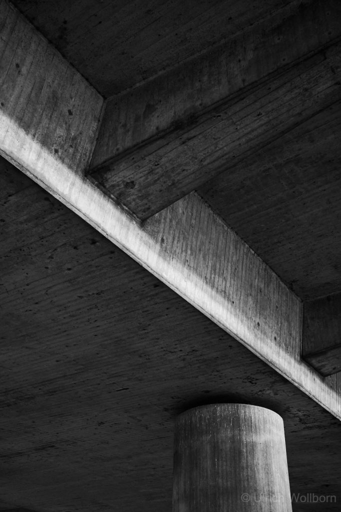 A black and white photo showing the underside of a concrete structure with beams and a cylindrical pillar. The composition emphasizes geometric shapes, textures, and the interplay of light and shadow in urban architecture.