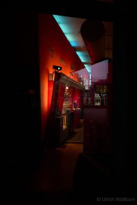 Dark bar with red wall illuminated by warm light, featuring glasses and bottles, reflected in a glass pane.